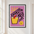 A framed art print featuring a retro-style illustration of an Amaretto Sour cocktail on a pink background, hung on a white wall next to a window.