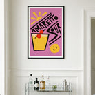 A framed art print featuring a retro 'Amaretto Sour' cocktail design in pink, yellow, black, and red, displayed on a white wall above a glass bar cart with bottles and glasses.