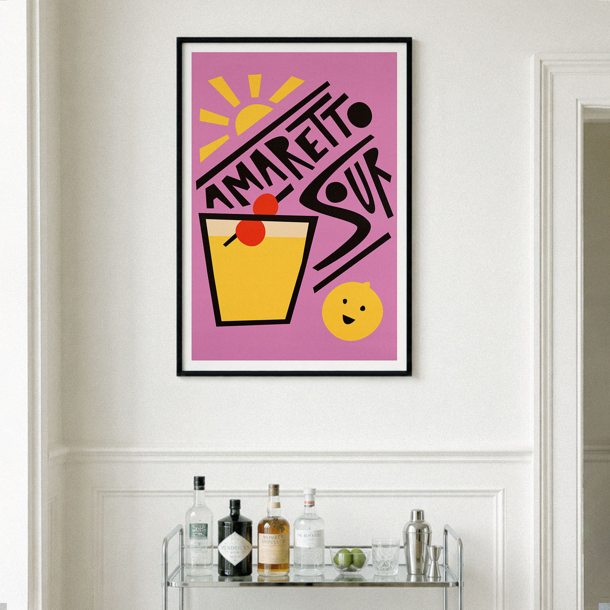 A framed art print featuring a retro 'Amaretto Sour' cocktail design in pink, yellow, black, and red, displayed on a white wall above a glass bar cart with bottles and glasses.