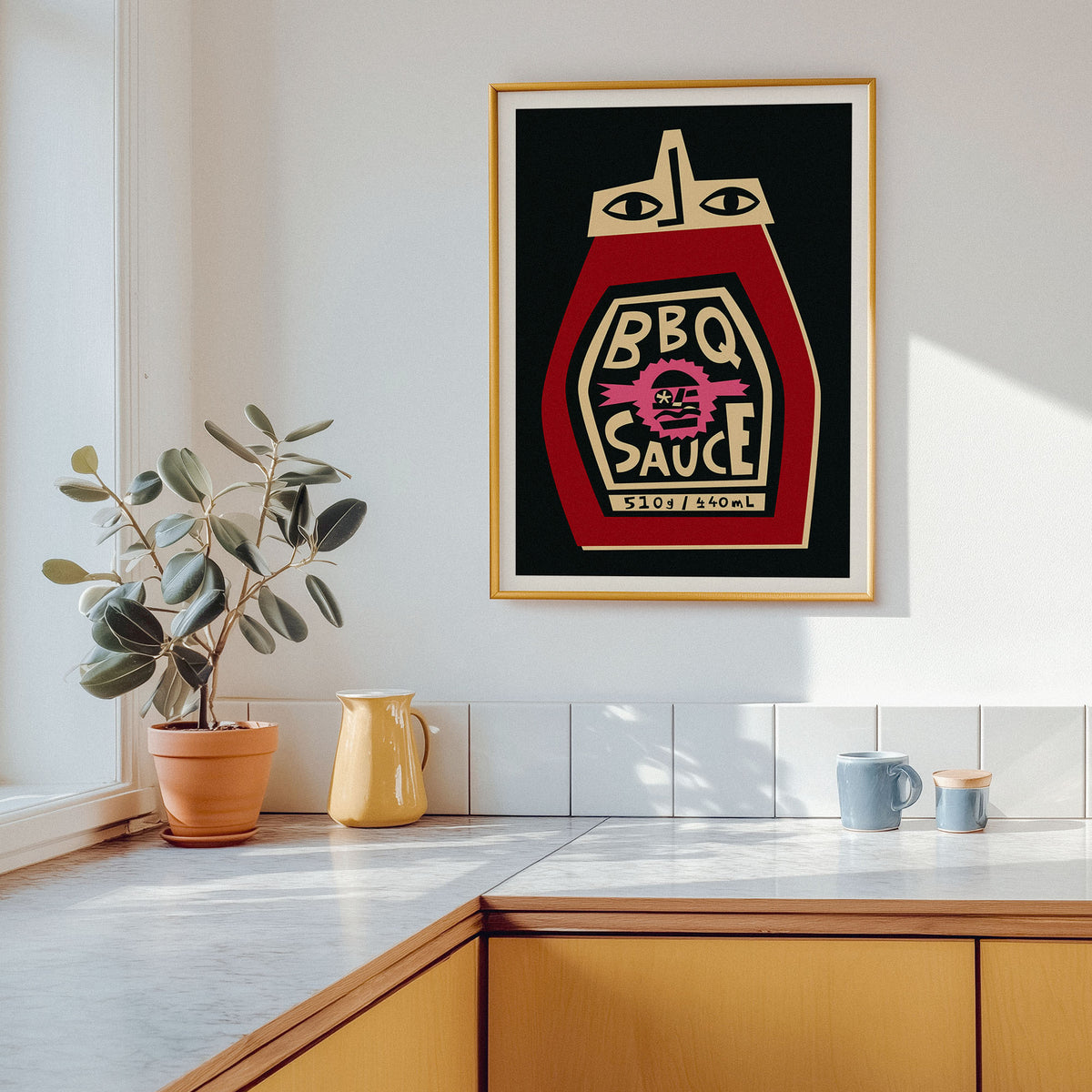 A framed art print featuring a stylized ketchup bottle labeled 'BBQ SAUCE' hangs on a white kitchen wall above a countertop with a potted plant and mugs.