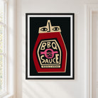 A framed art print of a stylized ketchup bottle labeled 'BBQ SAUCE' hanging on a white wall next to a window.