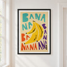 A framed art print featuring a retro-style illustration of three bananas and the word 'BANANA' repeated in colorful letters, hanging on a white wall next to a window.