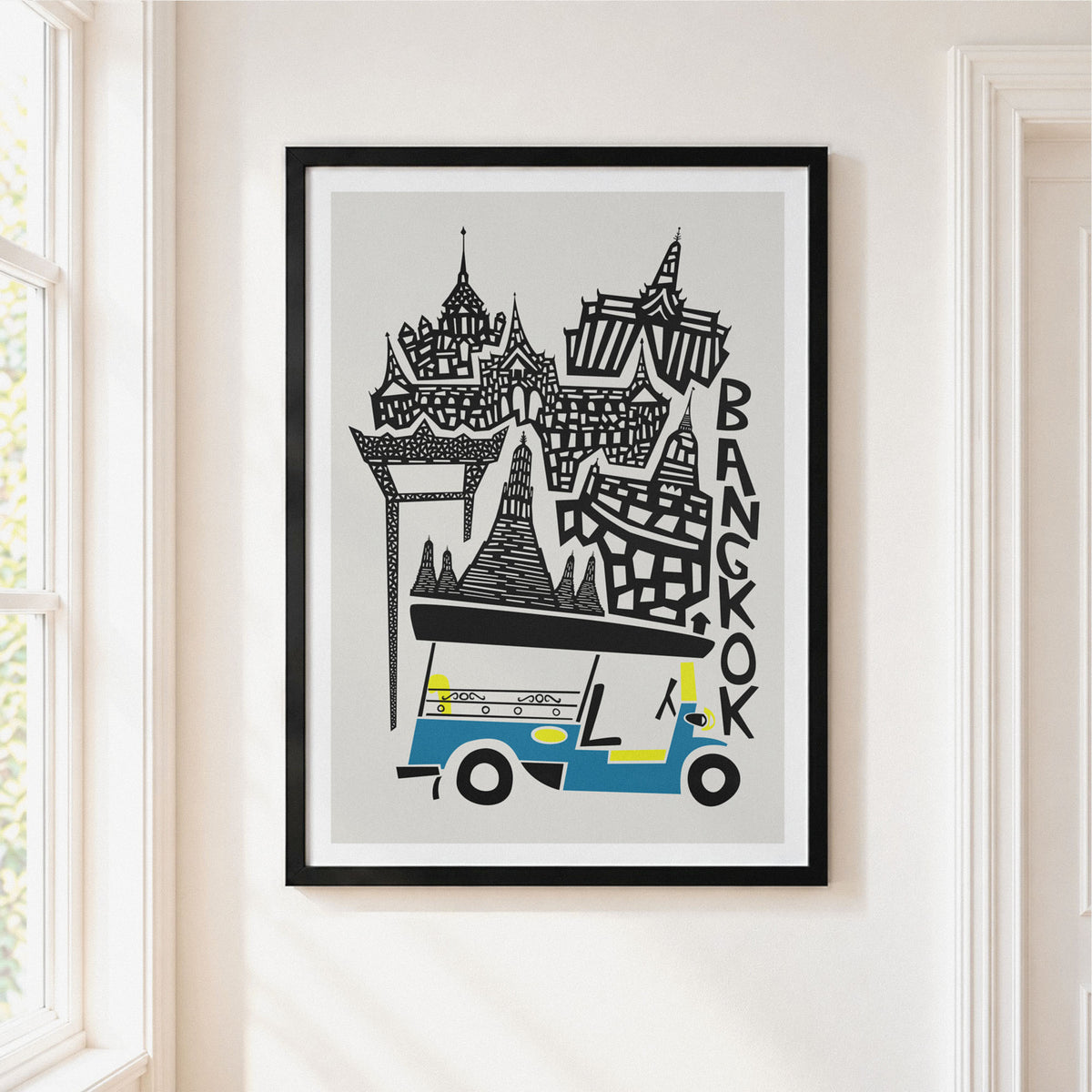 A framed art print featuring a stylized black and white illustration of Bangkok landmarks and a blue-and-yellow tuk-tuk, displayed on a light beige wall next to a window.
