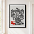 A framed art print featuring a stylized black and white illustration of Barcelona landmarks with a red cable car and the word 'BARCELONA' at the bottom, displayed on a beige wall next to a window.