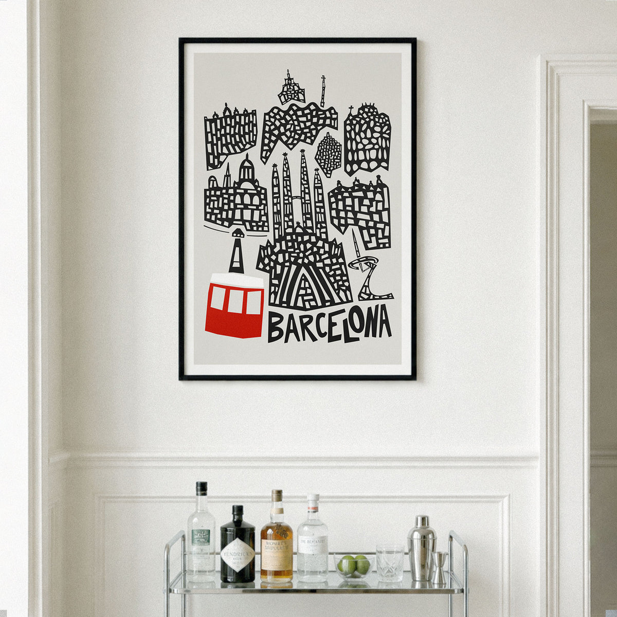 A framed black and white travel poster of Barcelona with a red cable car illustration, displayed on a white wall above a glass shelf with bottles.