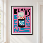 A framed art print featuring a bold graphic design with the words 'BEANS TOOT' and a stylized bean jar illustration, hung on a white wall next to a window.