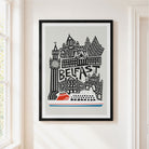 A framed art print featuring a stylized black and white illustration of Belfast landmarks with a red and white ferry at the bottom, hanging on a light beige wall next to a window.