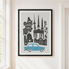 A framed art print featuring a stylized black line drawing of Berlin landmarks and a light blue car on a light gray background, hanging on a cream-colored wall next to a window.