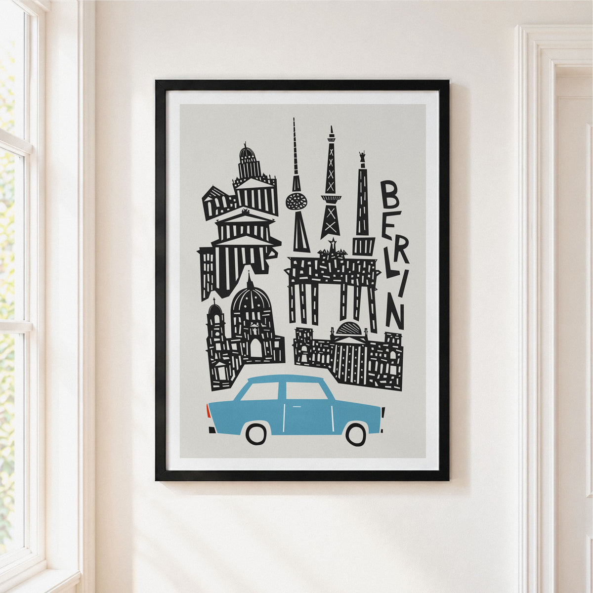 A framed art print featuring a stylized black line drawing of Berlin landmarks and a light blue car on a light gray background, hanging on a cream-colored wall next to a window.