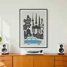 A framed art print featuring a stylized black line drawing of Berlin landmarks and a light blue car on a light gray background, hanging on a white wall above a wooden credenza with speakers and a record player.