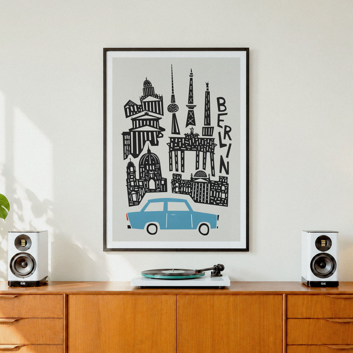 A framed art print featuring a stylized black line drawing of Berlin landmarks and a light blue car on a light gray background, hanging on a white wall above a wooden credenza with speakers and a record player.