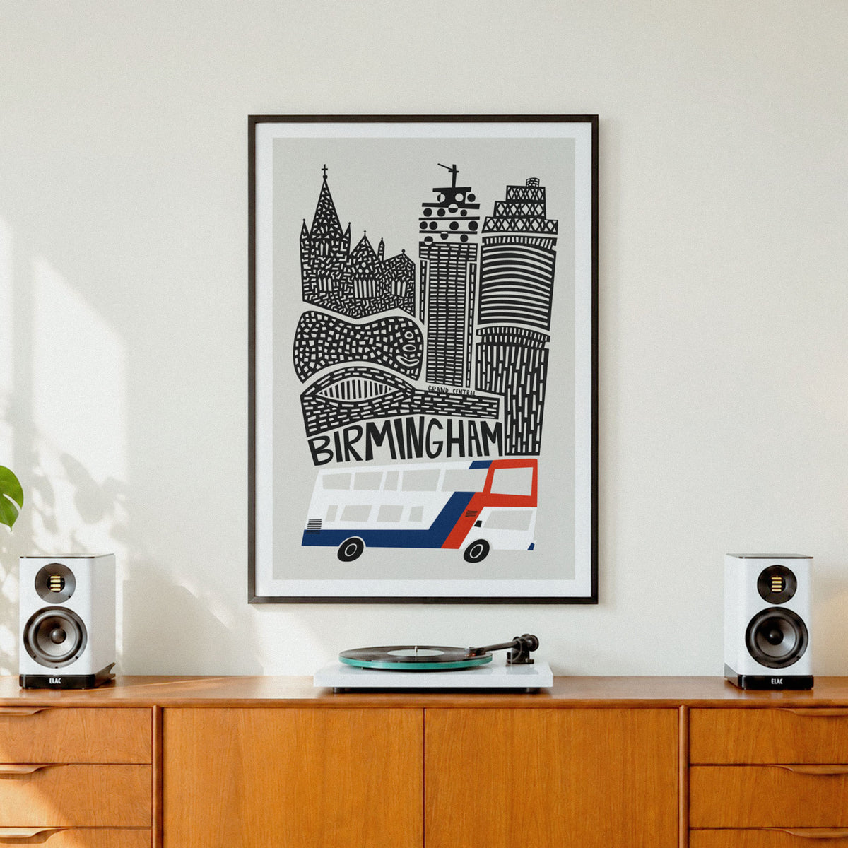 A framed black and white London-style cityscape art print with the word BIRMINGHAM and a red-and-white bus, hanging on a light wall above a wooden cabinet with speakers and a record player.