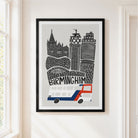A framed art print featuring a stylized black and white illustration of the Birmingham skyline with a red and blue double-decker bus, hanging on a light beige wall next to a window.