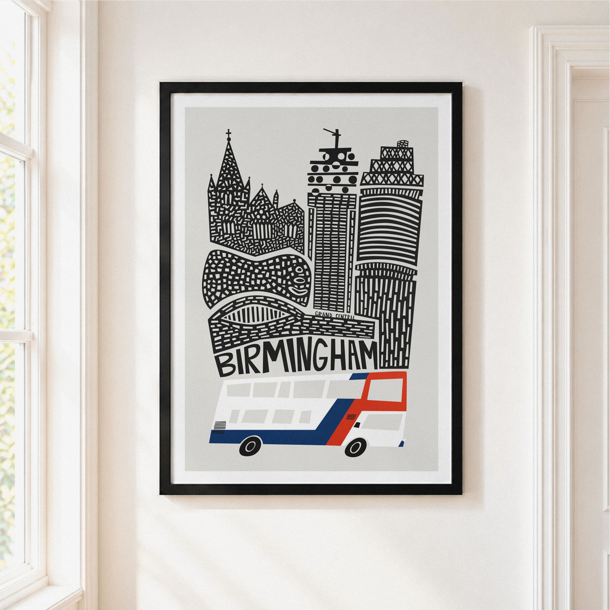 A framed art print featuring a stylized black and white illustration of the Birmingham skyline with a red and blue double-decker bus, hanging on a light beige wall next to a window.