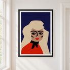 A framed minimalist art print of a stylized woman with blonde hair, a red top, and a black bow against a dark blue background, hanging on a white wall next to a window.