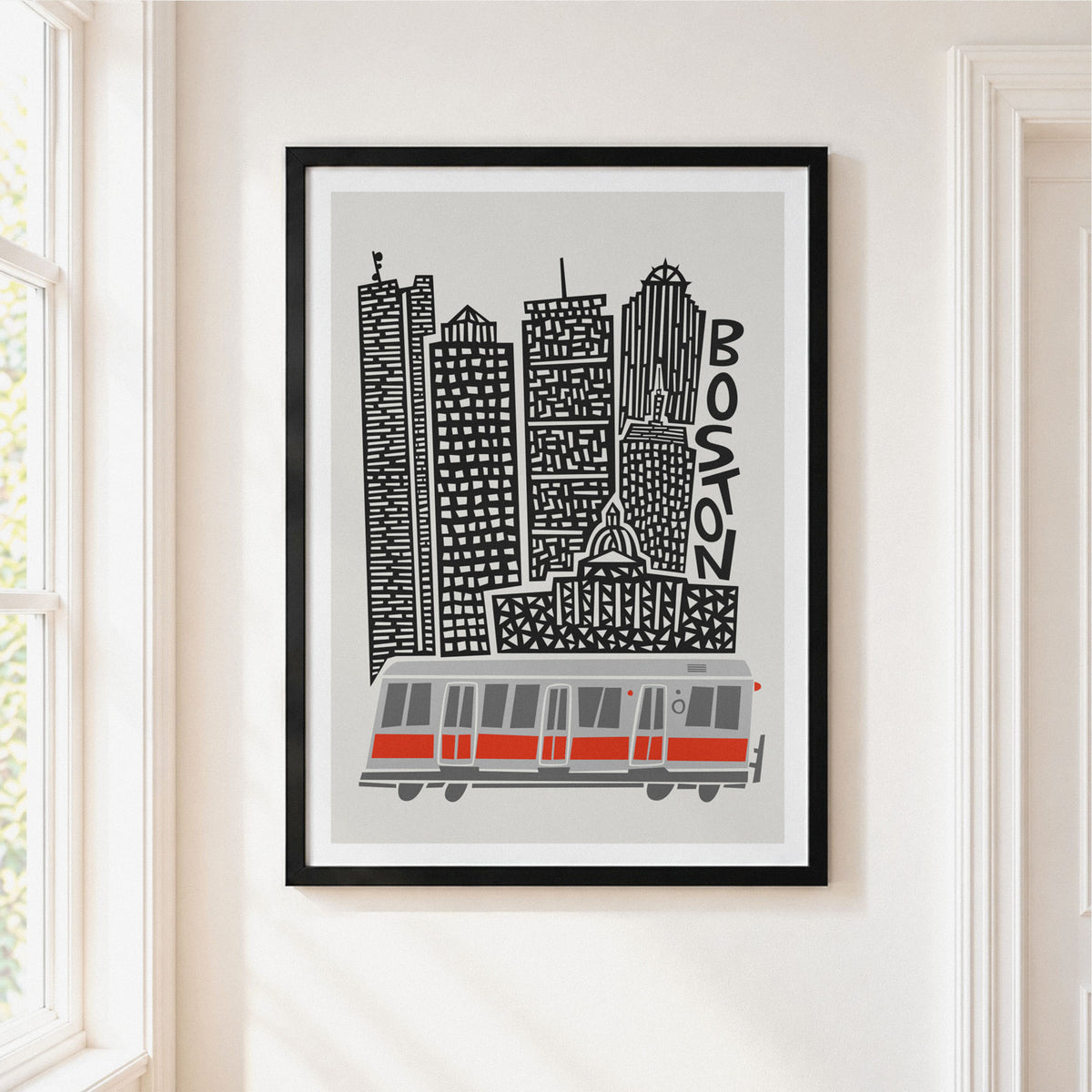 A framed art print featuring a stylized black and white Boston skyline with a red and gray cable car, hanging on a white wall next to a window.