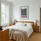 A styled bedroom interior featuring a wooden bed with white bedding and a beige linen throw, flanked by matching wooden nightstands with lamps. A framed black and white Boston cityscape print hangs on the white wall above the bed.