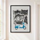 A framed art print featuring a stylized black and white illustration of Brighton landmarks with a blue scooter at the bottom, displayed on a cream-colored wall next to a window.