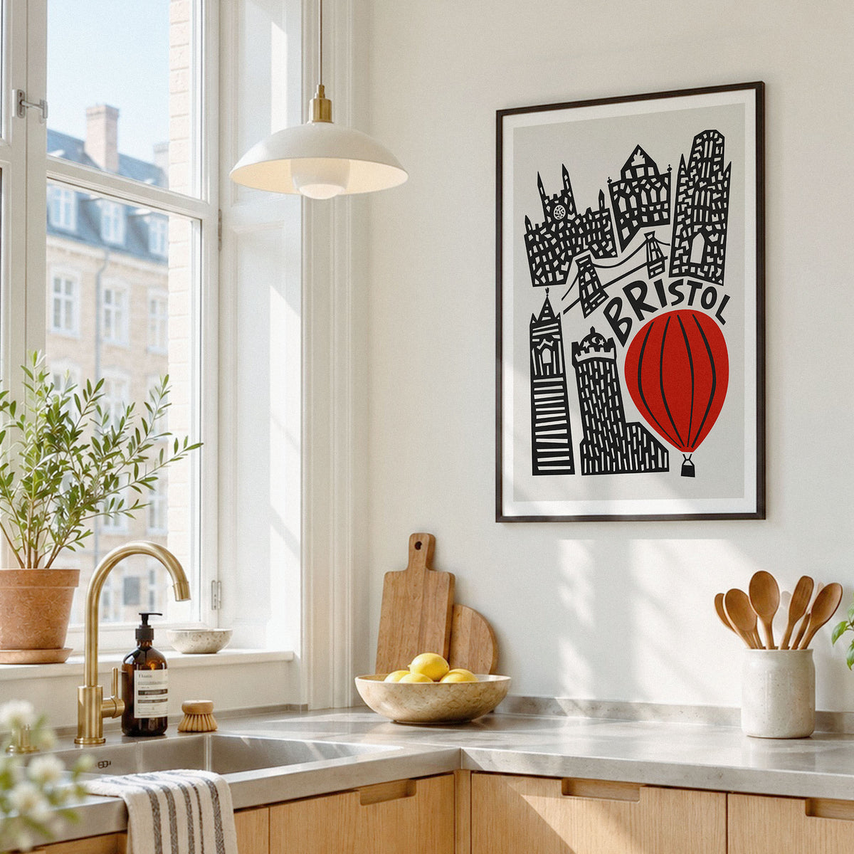 A framed art print featuring a black and white illustrated city skyline with a red hot air balloon and the word 'Bristol' is displayed on a white wall in a bright, modern kitchen.
