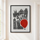 A framed art print featuring a stylized black and white illustration of Bristol landmarks and a red hot air balloon, hanging on a light beige wall next to a window.