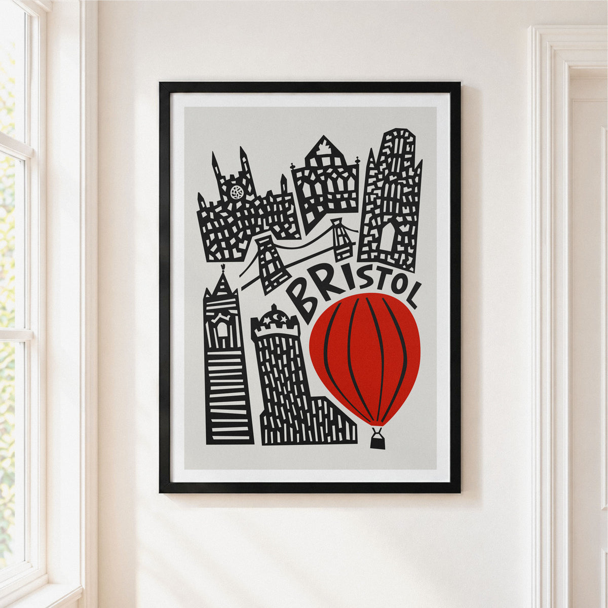 A framed art print featuring a stylized black and white illustration of Bristol landmarks and a red hot air balloon, hanging on a light beige wall next to a window.