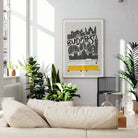 A framed Budapest cityscape art print hangs on a light gray wall above a beige sofa in a bright, modern living room with potted plants and natural sunlight.