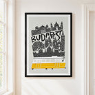 A framed Budapest travel art print hanging on a cream-colored wall next to a window. The poster features a black and white vector illustration of the Budapest skyline with the word 'BUDAPEST' written across it, and a yellow and white tram at the bottom.