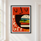 A framed retro-style poster featuring a stylized burger illustration with the words 'BURGER YUM' on a warm red-orange background, hung on a white wall next to a window.