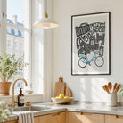 A framed black and white Cambridge cityscape art print with a blue bicycle illustration, hanging on a light wall in a bright, modern kitchen.