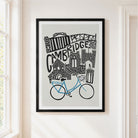 A framed art print featuring a black and white hand-drawn illustration of Cambridge landmarks with a blue bicycle, hanging on a warm off-white wall next to a window.