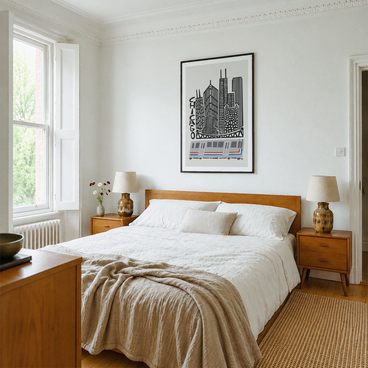 A styled bedroom interior featuring a wooden bed with white bedding and a beige throw blanket, flanked by matching wooden nightstands with lamps. A black and white framed cityscape print of Chicago hangs on the white wall above the bed.