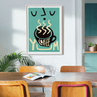 A framed art print featuring a stylized coffee cup with steam and the word 'COFFEE' in cream on a teal background, hanging on a light gray wall above a wooden dining table with chairs in a modern kitchen setting.