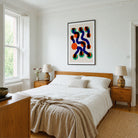 A modern bedroom featuring a colorful abstract wall art print with wavy organic shapes in navy, orange, red, and green on a cream background. The print is displayed above a wooden bed frame with white bedding and a beige linen throw.