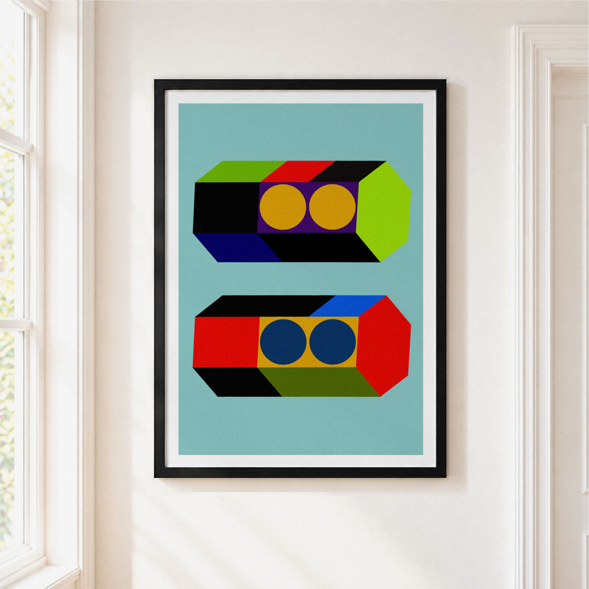 A framed abstract geometric art print hangs on a white wall in a bright room. The print features two stylized rectangular forms with colorful blocks and circles on a light blue background, enclosed in a black frame with a white mat.