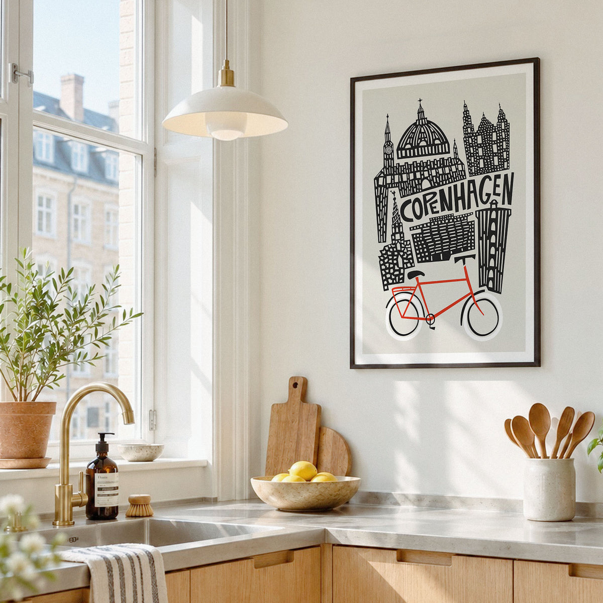 A framed art print featuring a black and white illustration of Copenhagen landmarks and a red bicycle, hanging on a light wall in a bright, modern kitchen.