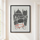 A framed art print featuring a stylized black line drawing of Copenhagen landmarks and a bright red bicycle on a light gray background, hanging on a white wall next to a window.
