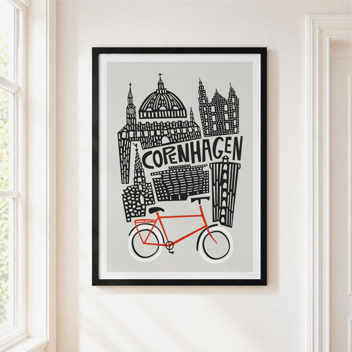 A framed art print featuring a stylized black line drawing of Copenhagen landmarks and a bright red bicycle on a light gray background, hanging on a white wall next to a window.
