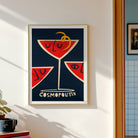 A framed art print featuring a stylized red cocktail glass with closed eyes on a dark navy background, hung on a white wall in a sunlit room. The print includes the word 'COSMOPOLITAN' at the bottom.