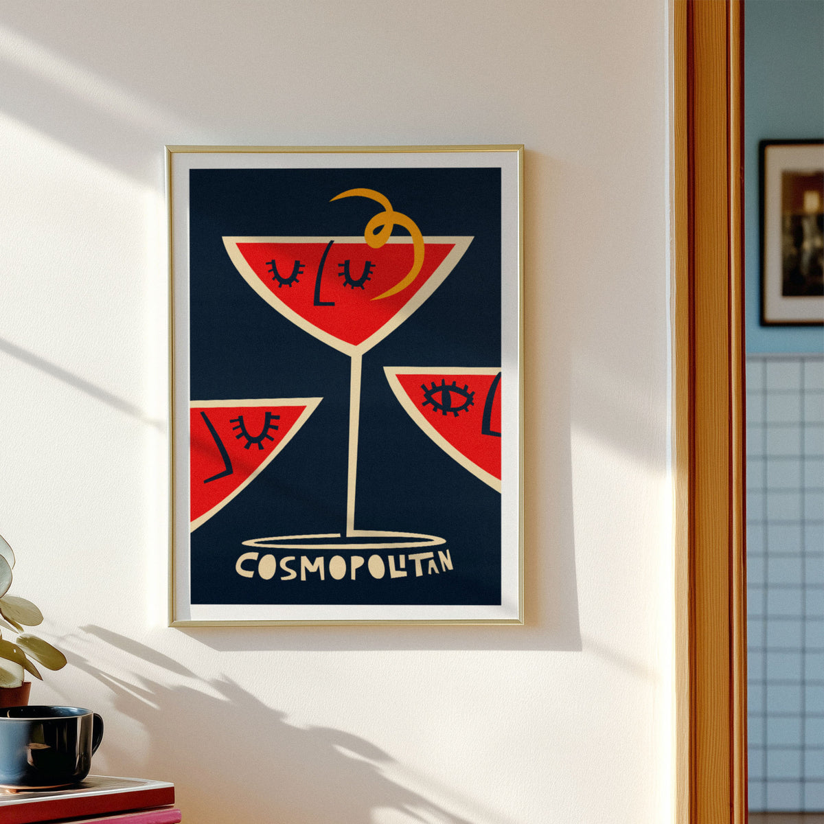 A framed art print featuring a stylized red cocktail glass with closed eyes on a dark navy background, hung on a white wall in a sunlit room. The print includes the word 'COSMOPOLITAN' at the bottom.