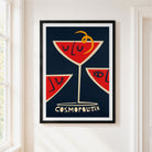 A framed art print featuring a stylized 'Cosmopolitan' cocktail design with red martini glass slices, cream details, and a yellow curl on a dark navy background, hung on a white wall next to a window.