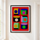A framed art print featuring a bold pop-art design of six colorful 3D cylinders on a red background, hung on a white wall in a bright room.