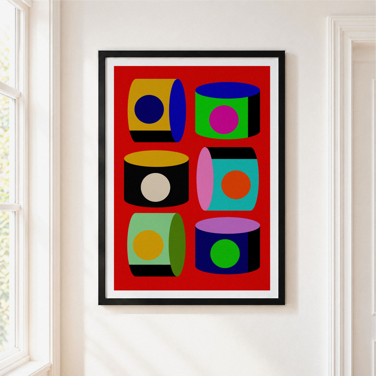 A framed art print featuring a bold pop-art design of six colorful 3D cylinders on a red background, hung on a white wall in a bright room.
