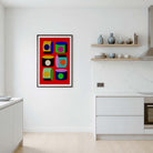 A framed abstract geometric art print with a bold red background and colorful square blocks hangs on a white wall in a modern kitchen setting.
