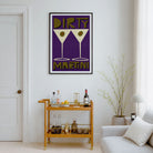 A framed wall art print featuring two martini glasses with the words 'DIRTY MARTINI' in olive green and cream on a purple background, displayed in a styled living room above a wooden bar cart.