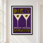 A framed art print featuring a retro  design with the words 'DIRTY MARTINI' and two martini glasses on a purple background, hanging on a white wall next to a window.