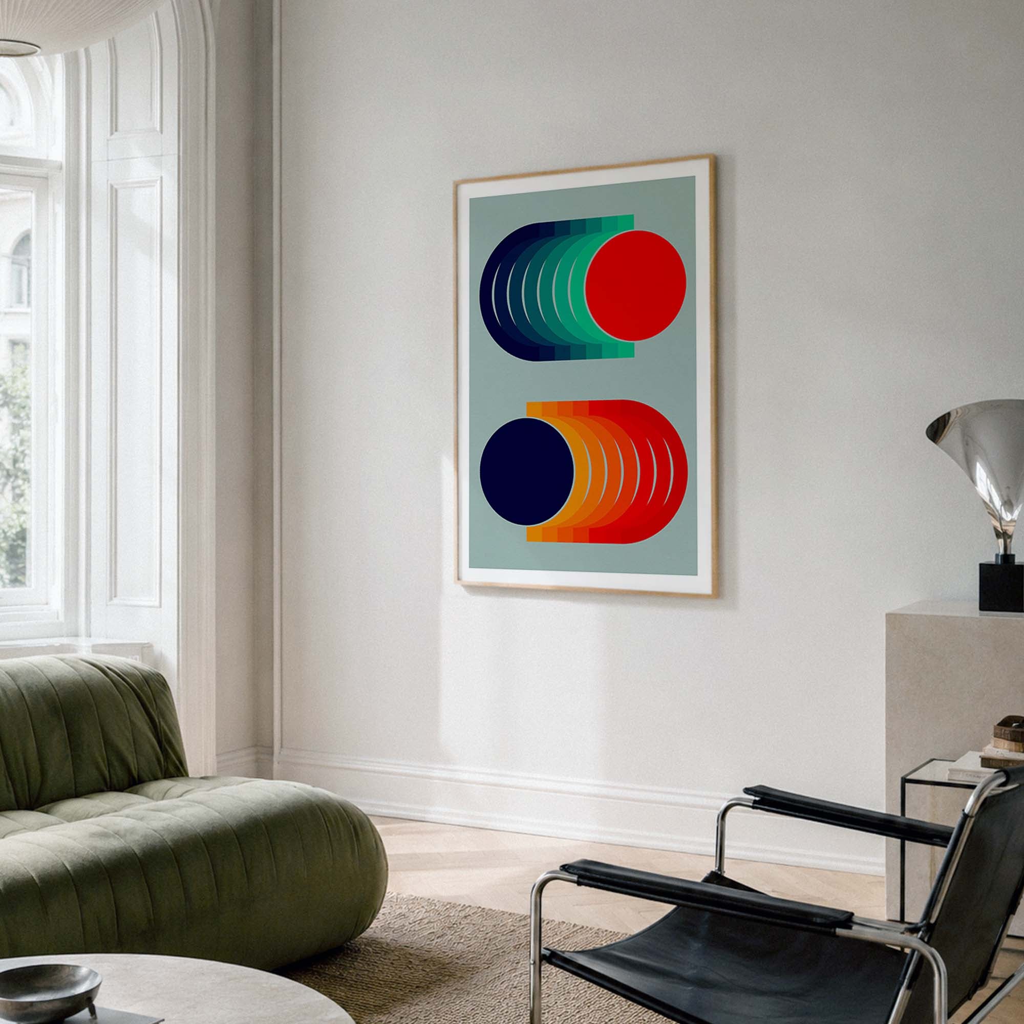 A framed abstract art print with geometric shapes in red, green, blue, and orange on a light gray background, hanging on a white wall in a modern living room setting.
