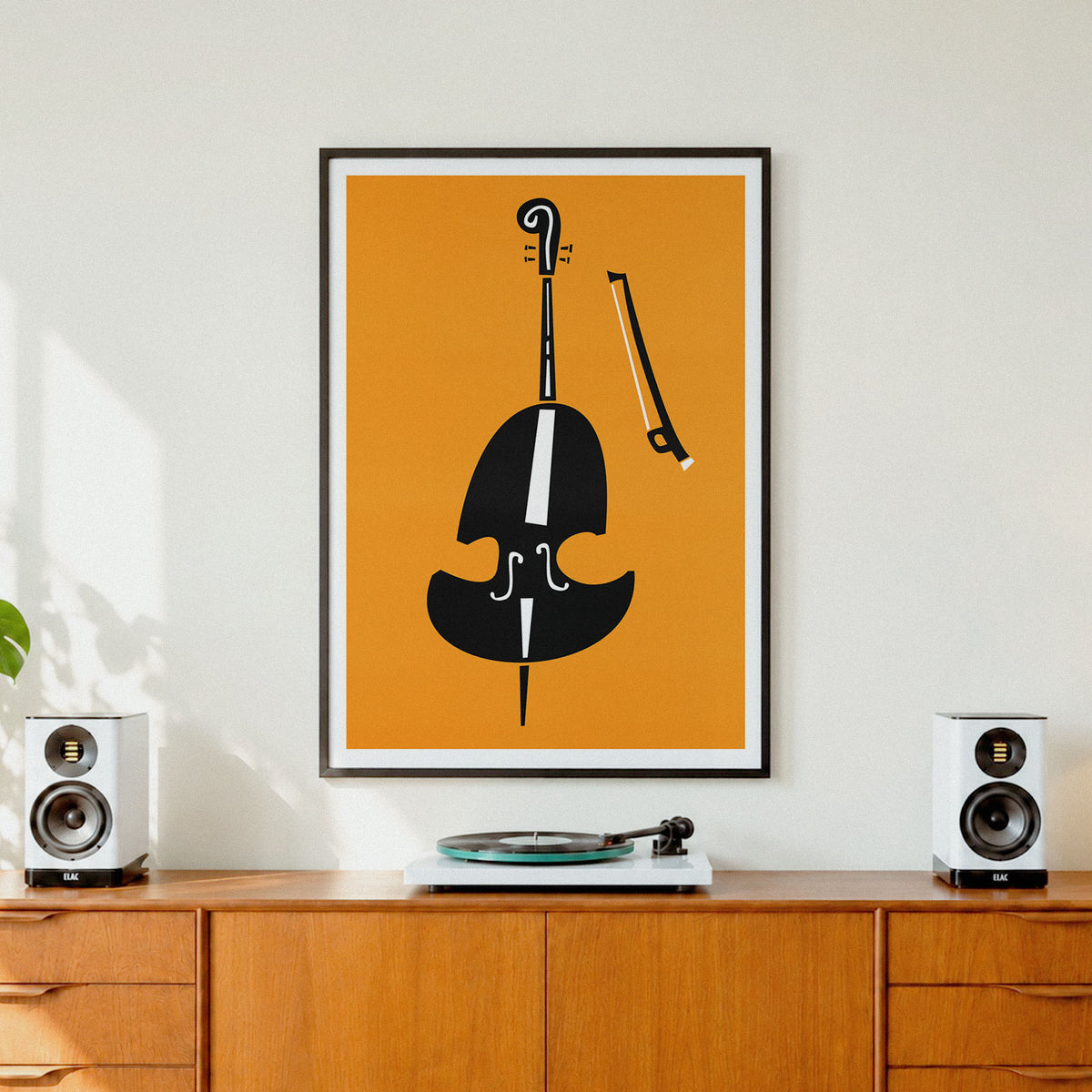 A framed minimalist art print featuring a black cello and bow on a mustard yellow background, displayed on a wooden cabinet in a modern room setting with speakers and a turntable.