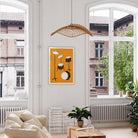 A framed art print featuring a minimalist orange, black, and white drum set illustration hangs on a white wall in a bright, modern living room with large windows, a rattan pendant light, and plants.
