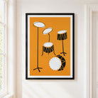 A framed minimalist drum set art print with an orange background, black and white drum illustrations, and a black frame, displayed on a white wall next to a window.