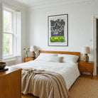 A styled bedroom interior with a wooden bed dressed in white bedding and a beige throw, flanked by matching wooden nightstands and lamps. A framed black-and-white typographic wall print of Dublin with a bright green train hangs above the bed.